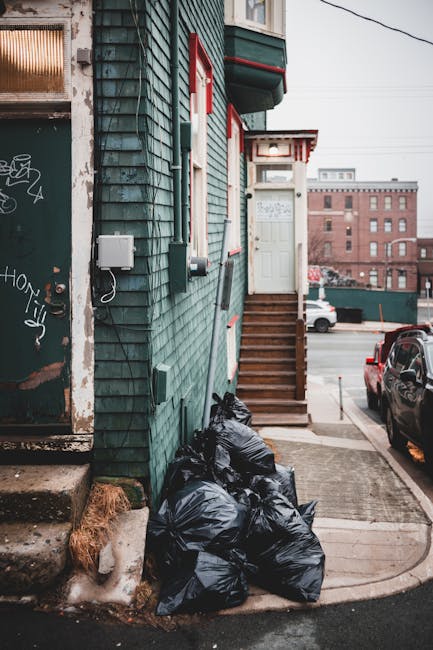 A collection of multiple black rubbish bags filled with waste, piled against the exterior green wooden wall of a residential building on a narrow street. The wall features horizontal wooden cladding painted in teal green, with some areas showing weathering and minor peeling. To the left, there is a graffiti-covered metal door with a small window and metal handle; a white electrical box is mounted on the wall nearby, with a loose cable hanging from it. A small set of wooden steps with a handrail leads up to a white front door situated under a small canopy with red trim, attached to the building's corner. The sidewalk is made of concrete, with a slightly raised curb. In the background, parked cars are visible along a street lined with other buildings, and overhead power lines cross the sky. The scene appears during daytime with soft, diffuse lighting typical of an overcast day, indicating an environment where private waste disposal or external rubbish collection may be taking place, as evidenced by the refuse accumulation outside the building. Waste Disposal Harrow often manages such rubbish and waste clearance in this area, supporting alternative waste handling for residents and businesses.