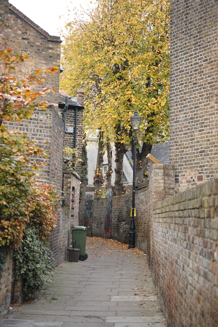 A collection of multiple black rubbish bags filled with waste, piled against the exterior green wooden wall of a residential building on a narrow street. The wall features horizontal wooden cladding painted in teal green, with some areas showing weathering and minor peeling. To the left, there is a graffiti-covered metal door with a small window and metal handle; a white electrical box is mounted on the wall nearby, with a loose cable hanging from it. A small set of wooden steps with a handrail leads up to a white front door situated under a small canopy with red trim, attached to the building's corner. The sidewalk is made of concrete, with a slightly raised curb. In the background, parked cars are visible along a street lined with other buildings, and overhead power lines cross the sky. The scene appears during daytime with soft, diffuse lighting typical of an overcast day, indicating an environment where private waste disposal or external rubbish collection may be taking place, as evidenced by the refuse accumulation outside the building. Waste Disposal Harrow often manages such rubbish and waste clearance in this area, supporting alternative waste handling for residents and businesses.