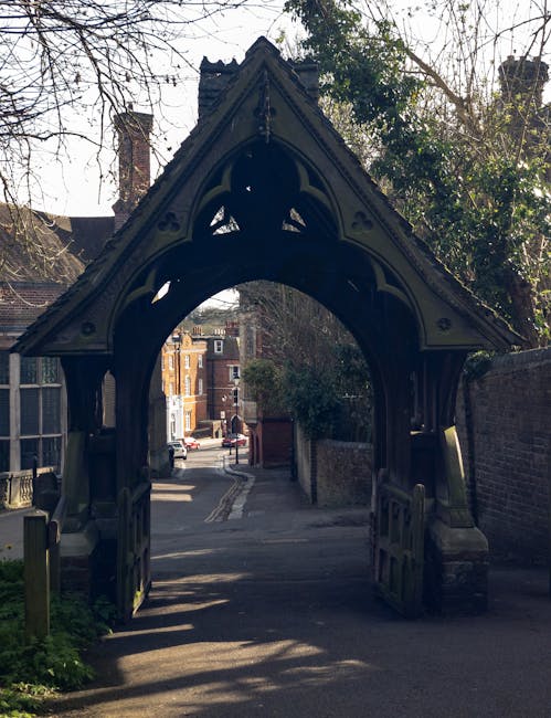The image depicts a historic archway with a pointed, Gothic-style timber frame featuring dark, weathered wood and ornate carved details, set against the backdrop of a residential street in Harrow. The arch's structure leans slightly to one side, with the timber beams glinting faintly in the daylight. The entrance opens onto a narrow paved lane bordered by brick walls and fences, with a glimpse of a street lined with traditional brick houses and parked cars in the distance. Shadows cast by nearby trees create a dappled pattern on the ground and the lower part of the arch. Surrounding foliage includes bare branches and some greenery, indicating early spring or late autumn. The environment suggests a quiet, residential area possibly requiring occasional private rubbish removal or on-site clearance, with the historic structure adding character to the setting. Waste Disposal Harrow could be involved in managing rubbish or waste collection from nearby properties, especially when local authority services are supplemented by independent rubbish removal solutions.