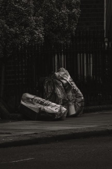 A collection of large black plastic rubbish bags, partially filled and slightly crumpled, are placed on the pavement near the edge of a residential area. The bags appear to contain garden or household waste, and some are tied at the top, showing signs of being ready for collection. They are positioned close to a black metal fence with vertical bars, behind which foliage from trees or shrubs is visible, providing a natural background. The scene is lit by natural daylight, with the overall atmosphere suggesting a routine outdoor waste collection or disposal process. The image reflects a typical private waste management setup, possibly managed by Waste Disposal Harrow for early delivery of rubbish from local residents or businesses, aligning with the context of alternative waste handling outside local authority services.