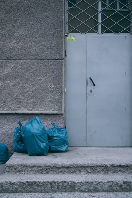 A group of four blue rubbish bags made from plastic, tied at the top and appearing to be filled with waste, are positioned on the concrete step outside a building's grey exterior wall, adjacent to a closed grey metal door with a black handle and lock. The wall has a textured finish, with a small yellow and green warning sticker near the top left corner of the door frame. Above the door, a section of diamond-patterned metal security grille covers a window, allowing some visibility through the glass. The setting appears to be an urban or commercial area, with the rubbish bags placed for collection or disposal, illustrating an example of private waste management that Waste Disposal Harrow might facilitate, especially for business premises seeking independent rubbish removal solutions. The surrounding environment is clean, with natural light illuminating the scene, emphasizing the practical aspect of rubbish collection outside a building entrance, relevant to the company's services in the West Harrow Station area.