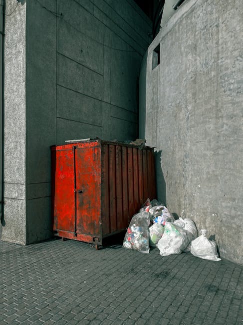 A collection of large black plastic rubbish bags, partially filled and slightly crumpled, are placed on the pavement near the edge of a residential area. The bags appear to contain garden or household waste, and some are tied at the top, showing signs of being ready for collection. They are positioned close to a black metal fence with vertical bars, behind which foliage from trees or shrubs is visible, providing a natural background. The scene is lit by natural daylight, with the overall atmosphere suggesting a routine outdoor waste collection or disposal process. The image reflects a typical private waste management setup, possibly managed by Waste Disposal Harrow for early delivery of rubbish from local residents or businesses, aligning with the context of alternative waste handling outside local authority services.