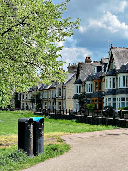 The image depicts a row of traditional Victorian-style terraced houses with bay windows, some with decorative wooden framing, situated along a quiet residential street. In the foreground, there are two black waste bins positioned on a grassy verge adjacent to a paved footpath. The bins are cylindrical with slightly domed lids and are placed side by side close to each other, with one slightly taller than the other. To the left, a large tree with vibrant green leaves extends its branches over the scene, partially shading the grassy area and the bins. The homes have brick facades with some featuring light-colored stone accents, chimneys on the rooftops, and a mix of slate and tiled roofing. The background displays a partly cloudy sky with patches of blue, suggesting a clear and bright day. This scene, set in a suburban neighborhood, subtly relates to the topic of rubbish removal services by illustrating the typical environment where domestic waste collection and private disposal arrangements may take place, exemplifying the kind of setting Waste Disposal Harrow might operate within for garden waste clearance or general rubbish collection.