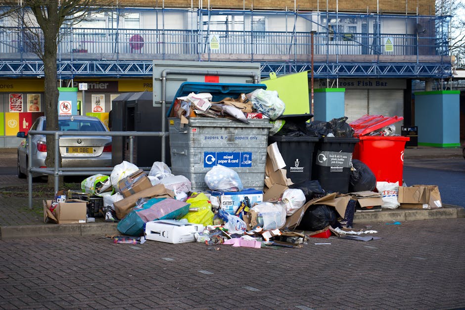 An outdoor scene showing a large assortment of overflowing rubbish and waste materials piled around a communal recycling bin on a paved area in front of a commercial building. The waste includes various cardboard boxes, black and green trash bags, some flattened and partially torn, mixed paper and plastic packaging, and a few discarded items such as a white appliance part and a yellow plastic container. The recycling bin, labeled for mixed paper and cardboard, is partially open with some waste spilling out, and is situated close to a black bin and a red waste receptacle, all placed behind a short metal railing. In the background, a building with a blue metal staircase and scaffolding, suggesting ongoing maintenance work, is visible along with storefronts with advertising posters. A parked silver car with a yellow registration plate is positioned to the left, next to a leafless tree on a pedestrian pavement. The scene is set during daytime with natural light illuminating the area, emphasizing the disorderly state of the waste, which could be indicative of issues related to private or improper rubbish management, as managed by waste disposal services like Waste Disposal Harrow.