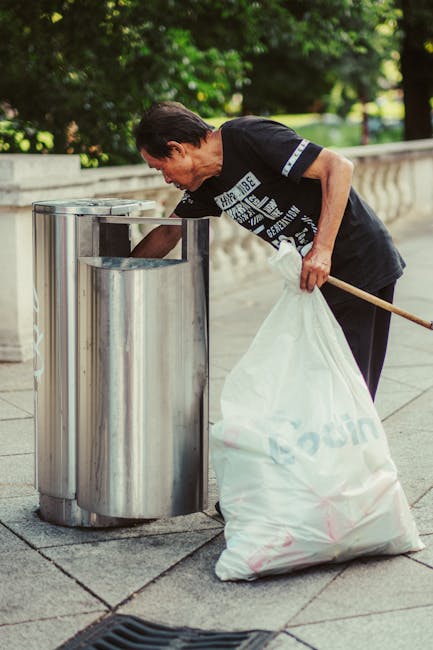 A middle-aged man with short dark hair, wearing a black T-shirt with white text and black trousers, is standing outdoors on a paved area near a stone railing. He is leaning forward, holding a large white plastic bag filled with rubbish in his left hand, while using a wooden stick or broom handle in his right hand to pick up or deposit waste into a modern, stainless steel public trash bin with a rounded top and multiple compartments. The background features lush green trees and foliage, suggesting a park or garden setting during daytime with natural lighting. The scene highlights public waste disposal activity, and the presence of the waste disposal company's service aligns with alternative rubbish removal or on-site waste collection practices relevant to private or community rubbish management.
