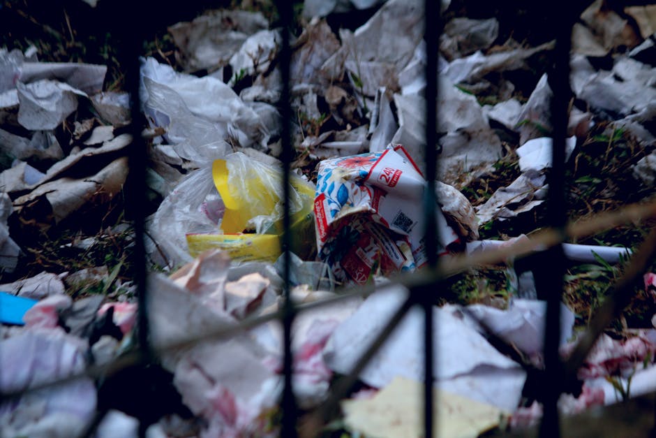 Close-up view of a small pile of discarded household waste scattered on the ground behind a black metal fence with vertical bars. The waste includes crumpled paper, plastic bags, and torn packaging, some with printed labels and bright colours such as yellow and red. The materials have varying textures, with the paper appearing wrinkled and the plastic bags shiny and smooth. The debris is spread unevenly across a muddy or earthy surface, with bits of grass and small twigs visible among the rubbish. Natural lighting highlights the different surfaces and the disorderly arrangement of the waste. The scene suggests an informal or unmanaged waste deposit, which could be subject to collection or disposal by services such as Waste Disposal Harrow for effective rubbish removal and proper environmental management.