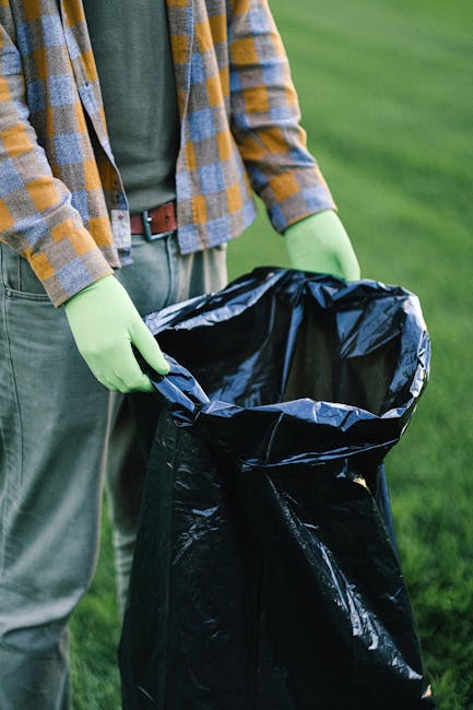 A black wheeled rubbish bin positioned outdoors on a grassy area with lush green foliage and shrubs in the background. The lid of the bin is closed, and the top is partially covered with a variety of garden waste, including dried grass, leaves, and small branches. The garden waste appears fresh and dry, with some of the weeds and stems protruding over the edges of the lid. To the left of the bin, there is a vibrant yellow flowering shrub, partially visible behind the green shrubbery, contributing to the natural environment setting. The scene is illuminated by natural sunlight, casting gentle shadows on the grass and highlighting the textures of the garden refuse and the surrounding plants. This scene illustrates a typical outdoor garden clearance, relevant to private waste handling services like those offered by Waste Disposal Harrow, supporting the context of alternative waste removal methods outside local authority collections.