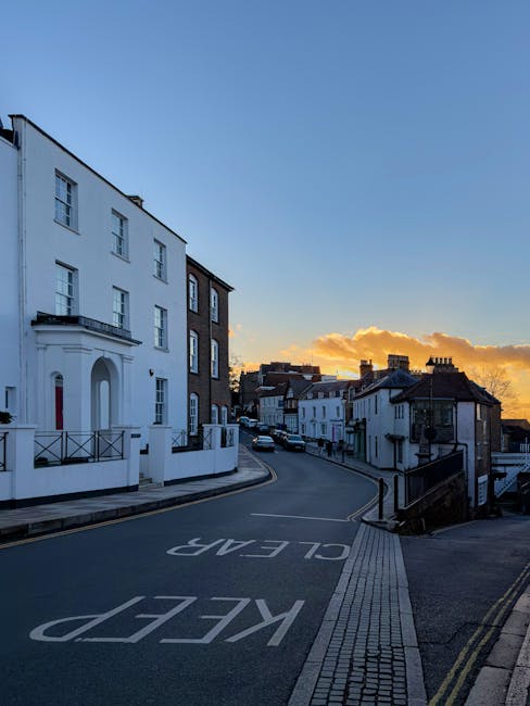 A quiet urban street scene during sunset showing a gently curving road with the words 'KEEP CLEAR' painted on the asphalt in large white letters. To the left, there is a white building with a smooth façade, prominent windows, and a small balcony, providing a contrast to the darker neighboring structure with a brick exterior. In the background, a row of terraced houses with sloped roofs and chimney stacks extends along the street. The street is bordered by a paved sidewalk with tactile paving strips next to the kerb, and there are a few parked cars visible further down the road. The sky above displays warm, golden hues from the setting sun, casting soft light over the scene, with some clouds illuminated in orange and yellow tones. This scene captures a typical residential area where private waste collection services like Waste Disposal Harrow could be involved in rubbish removal or on-site clearance activities, with the tidy street and clear markings emphasizing organized urban waste management practices.
