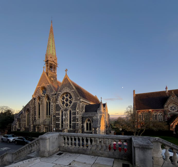A stone-paved balcony with a low, decorative balustrade overlooks a historic church constructed from dark flint and light stone, featuring intricate gothic-style detailing, pointed arches, rose windows, and a tall, slender spire with a greenish patina. The church is situated in a spacious outdoor area with surrounding trees and other historic buildings, illuminated by soft natural light at sunset or dawn. In the foreground, parked cars are visible along a driveway or street, and the scene appears quiet and well-maintained. The setting suggests a peaceful, urban environment where independent waste collection or rubbish removal services may occur nearby, with Waste Disposal Harrow potentially handling rubbish clearance from local properties or public spaces. The overall atmosphere emphasizes the clean, organized appearance of the area, suitable for professional waste management operations.
