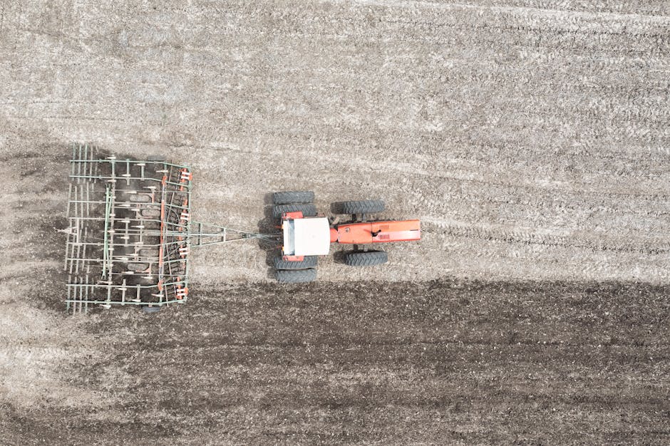 An overhead aerial view of a small orange tractor with black rubber tires, positioned in the centre of a large, freshly tilled field with dark, textured soil. Attached to the tractor's rear is a metal agricultural implement, resembling a specialised harrow or rake with multiple metal tines or bars, which appears to be used for soil cultivation or preparation. The background consists of expansive, uniform soil stretches without additional objects or vegetation, indicating an open rural environment. The lighting is natural, with soft, diffuse illumination, suggesting an overcast day. The scene reflects elements of land management or preparation suitable for agricultural activities and subtly aligns with themes of land clearing or site preparation services often associated with waste or debris clearance undertaken by companies like Waste Disposal Harrow, facilitating alternative site clean-up or on-site clearance processes.